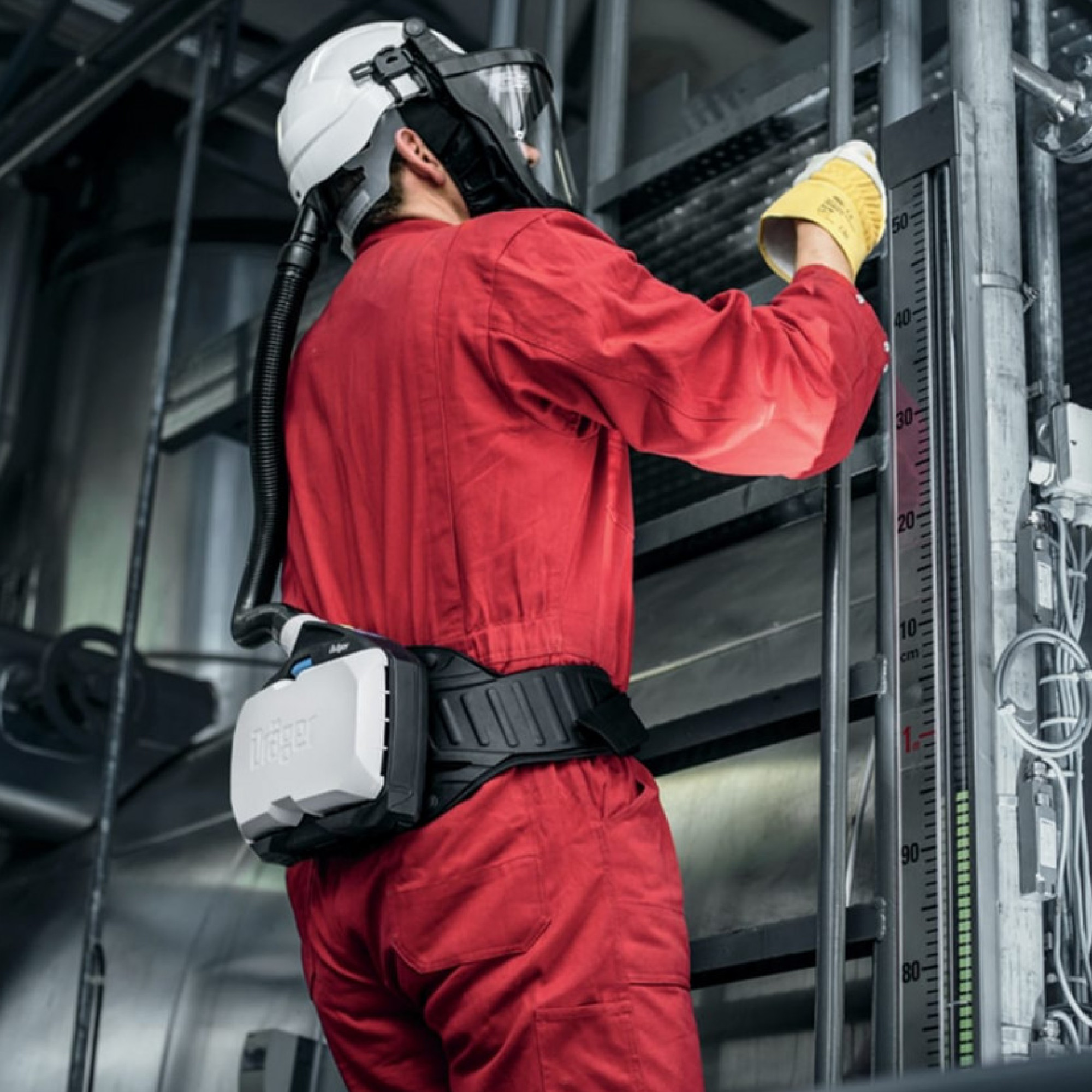 Ein Arbeiter in einem roten Overall steht auf einer Leiter und betrachtet ein Messgerät. Er trägt einen Schutzhelm mit Visier und ein Atemschutzgerät, das an einem Gurt befestigt ist. Die Umgebung wirkt industriell, mit verschiedenen Maschinen im Hintergrund.