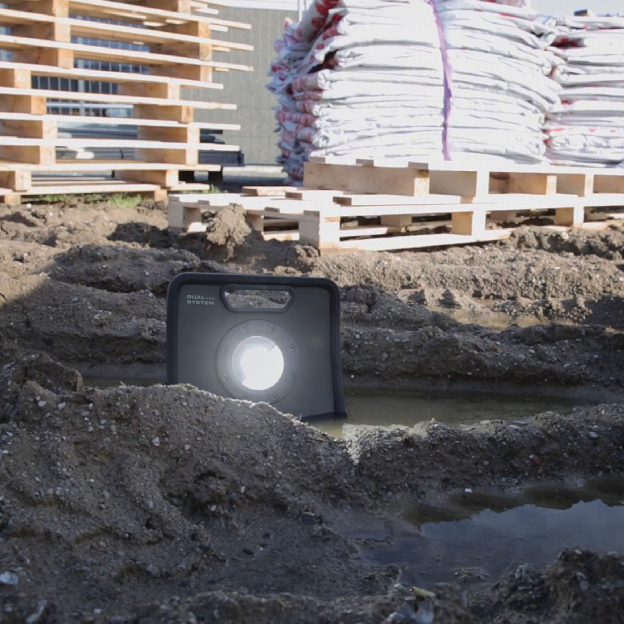 Ein tragbarer Baustrahler steht in einem trockenen Graben, umgeben von losem Sand und einer kleinen Wasserpfütze. Im Hintergrund sind Holzpaletten und Säcke, die scheinbar Baustellenmaterialien enthalten. Der Strahler ist eingeschaltet und strahlt ein helles Licht aus.