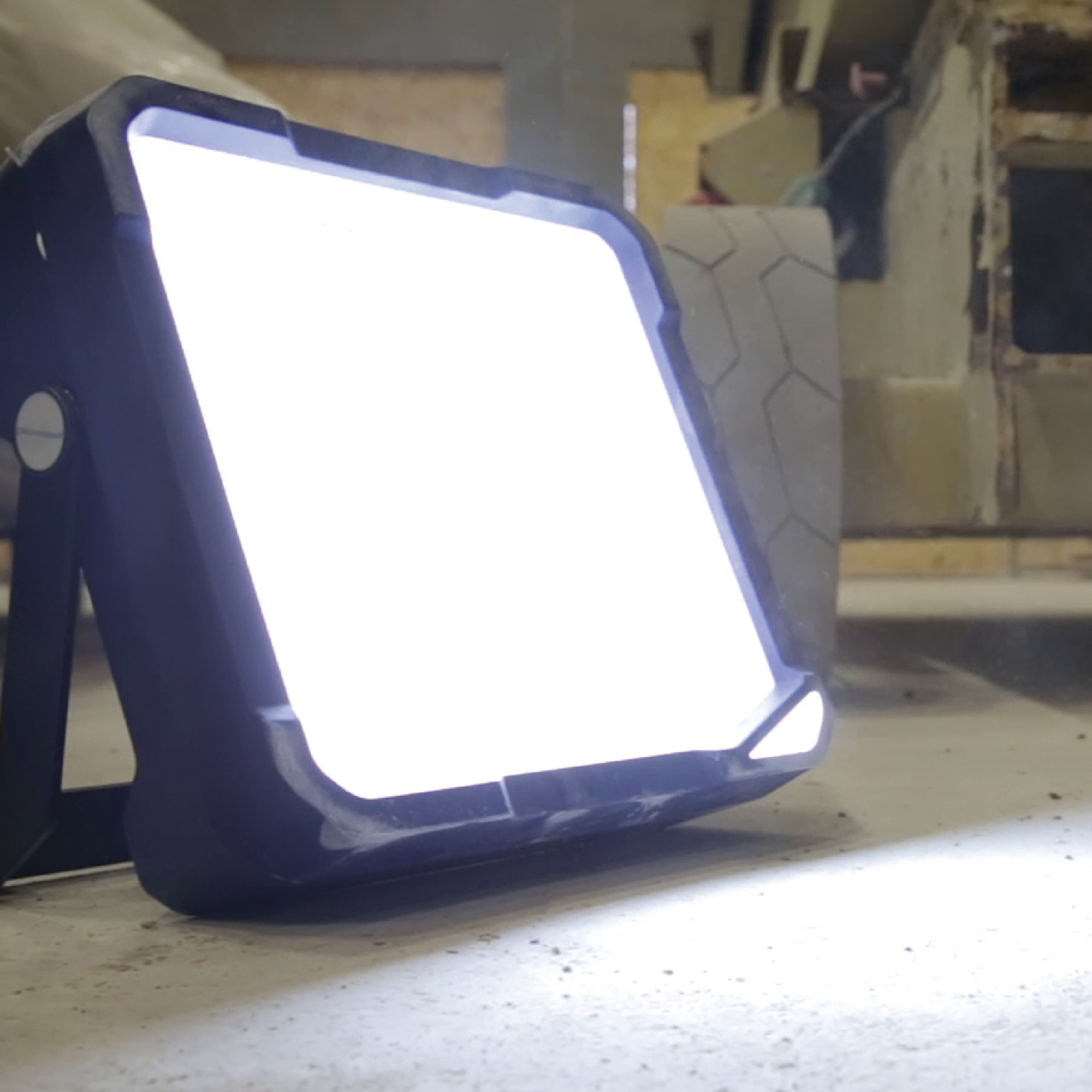 Eine helle Arbeitslampe steht auf einem Tisch und strahlt starkes Licht aus. Im Hintergrund ist ein unscharfes Objekt zu erkennen. Die Lampe hat einen stabilen Rahmen und eignet sich für die Beleuchtung von Arbeitsbereichen.