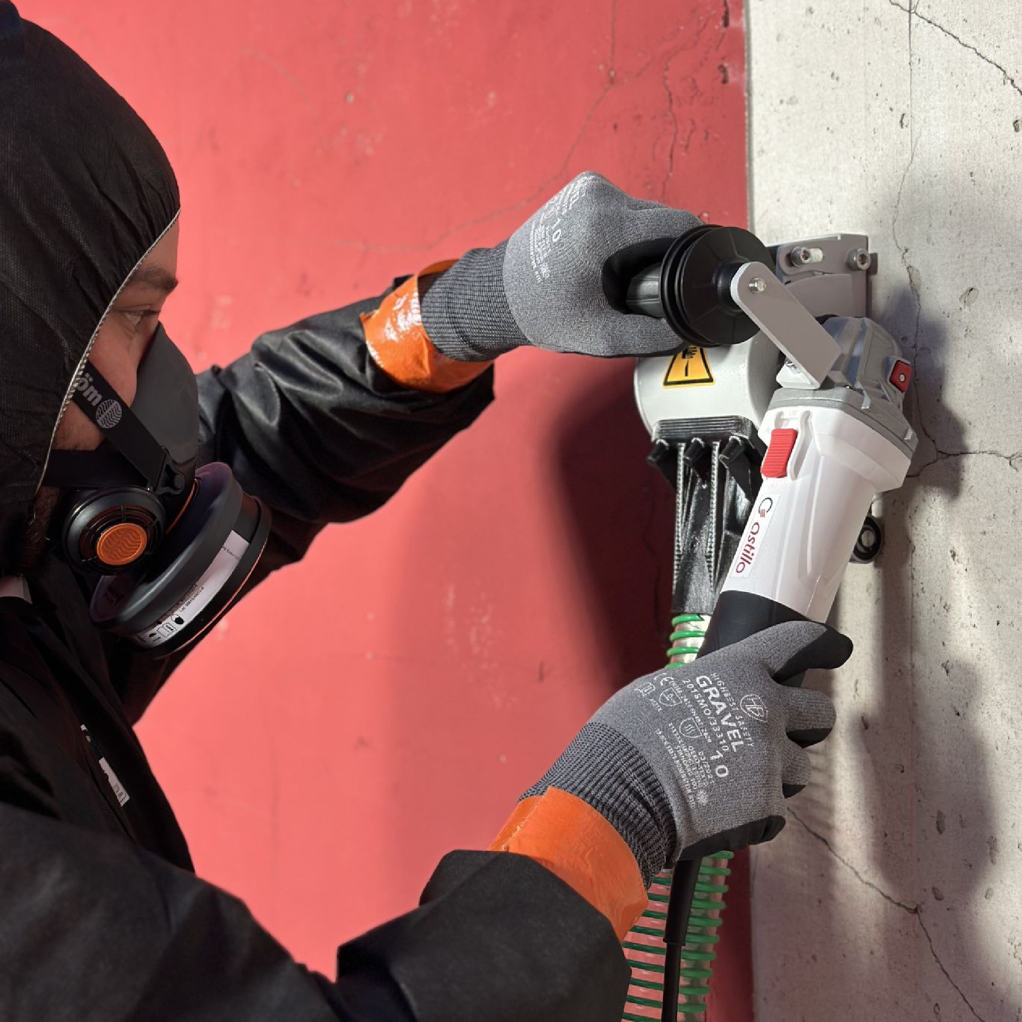 Ein Handwerker in Schutzausrüstung bedient eine Wandbearbeitungsmaschine an einer Betonkante. Der Hintergrund ist rot, und die Maschine wird mit beiden Händen festgehalten. Schutzmasken und spezielle Handschuhe sind sichtbar, was auf Sicherheitsmaßnahmen hinweist.