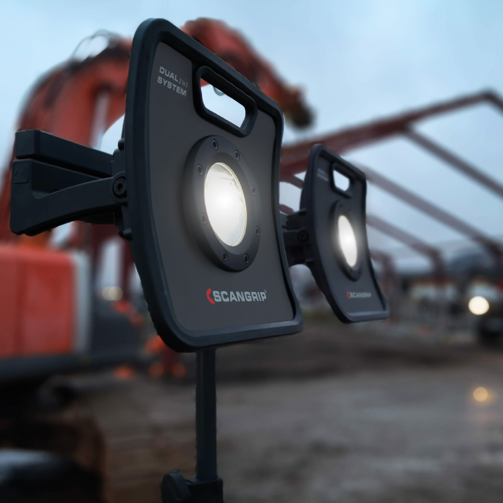 Das Bild zeigt zwei leistungsstarke LED-Arbeitslichter von SCANGRIP, montiert auf einem Stativ. Ihre hellen Lichter leuchten in einem grauen, dämmerigen Umfeld, das möglicherweise eine Baustelle darstellt. Der Hintergrund ist verschwommen, während die Lichter im Vordergrund klar erkennbar sind.