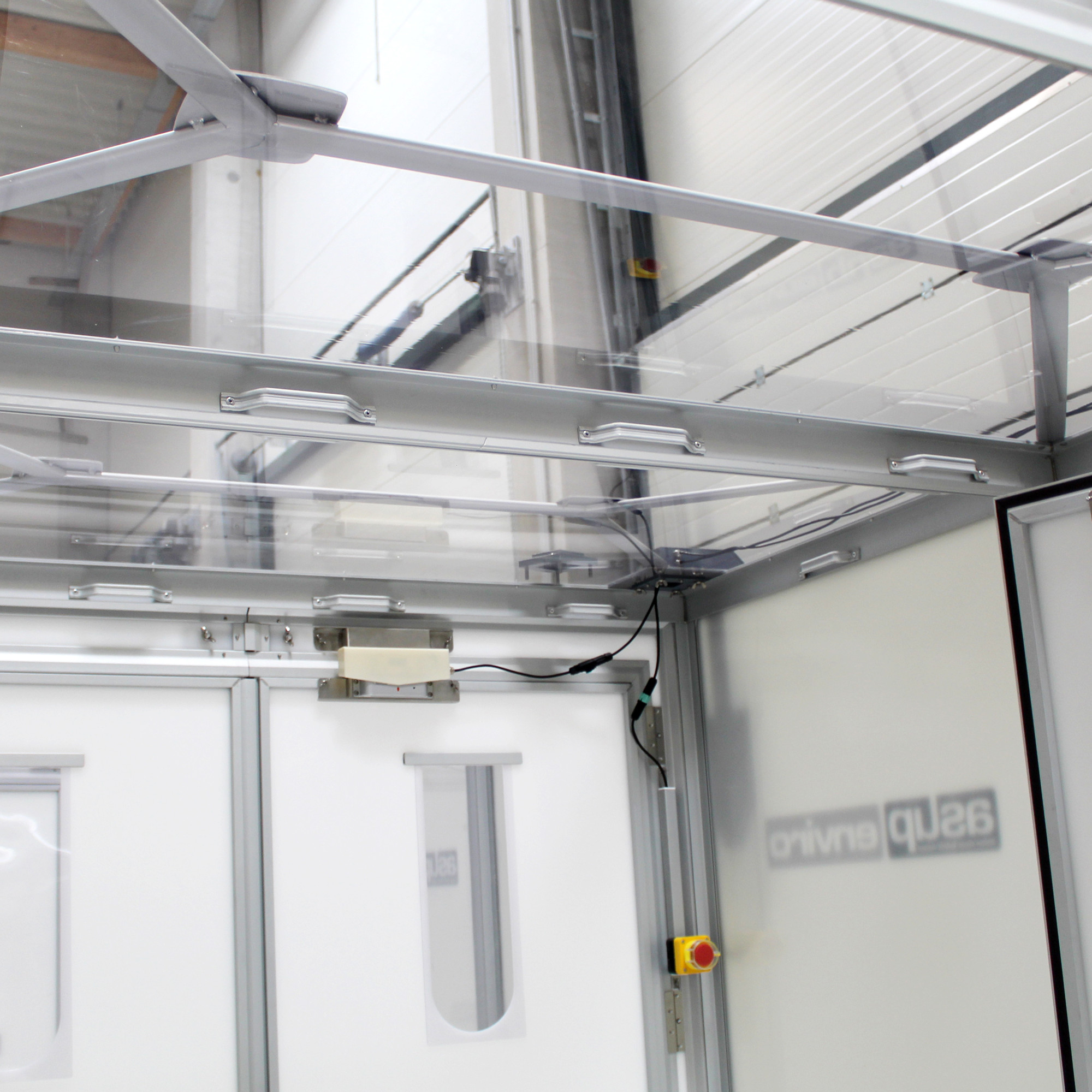 Innenraum einer 2-Kammer-Materialschleuse mit transparenter Decke, zwei Türen mit Sichtfenstern, Steuergerät, Wandkabeln und gelbem Bedienelement
