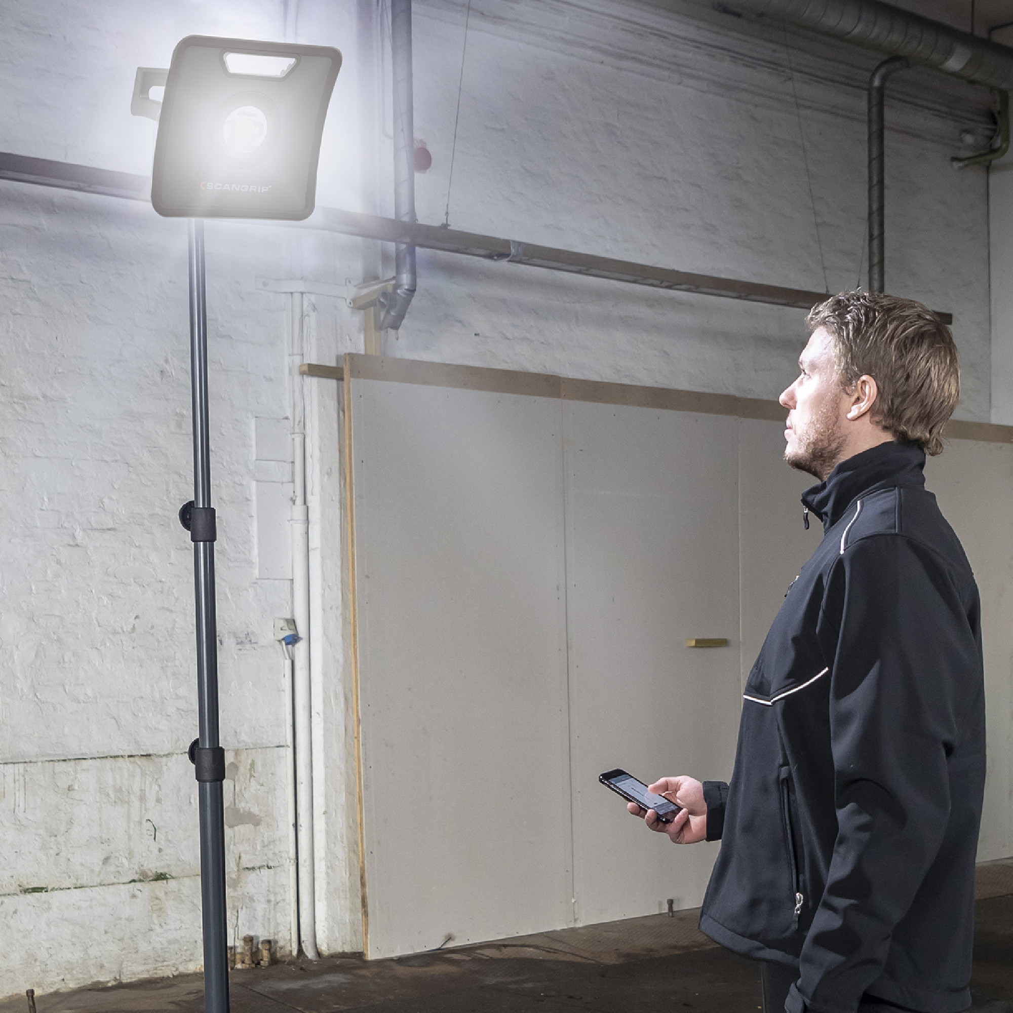 Ein Mann steht in einem Raum und schaut zu einem hellen Baustrahler auf einem Ständer. Er hält ein Smartphone in der Hand und trägt eine dunkle Jacke. Der Raum hat eine einfache, helle Wand im Hintergrund.
