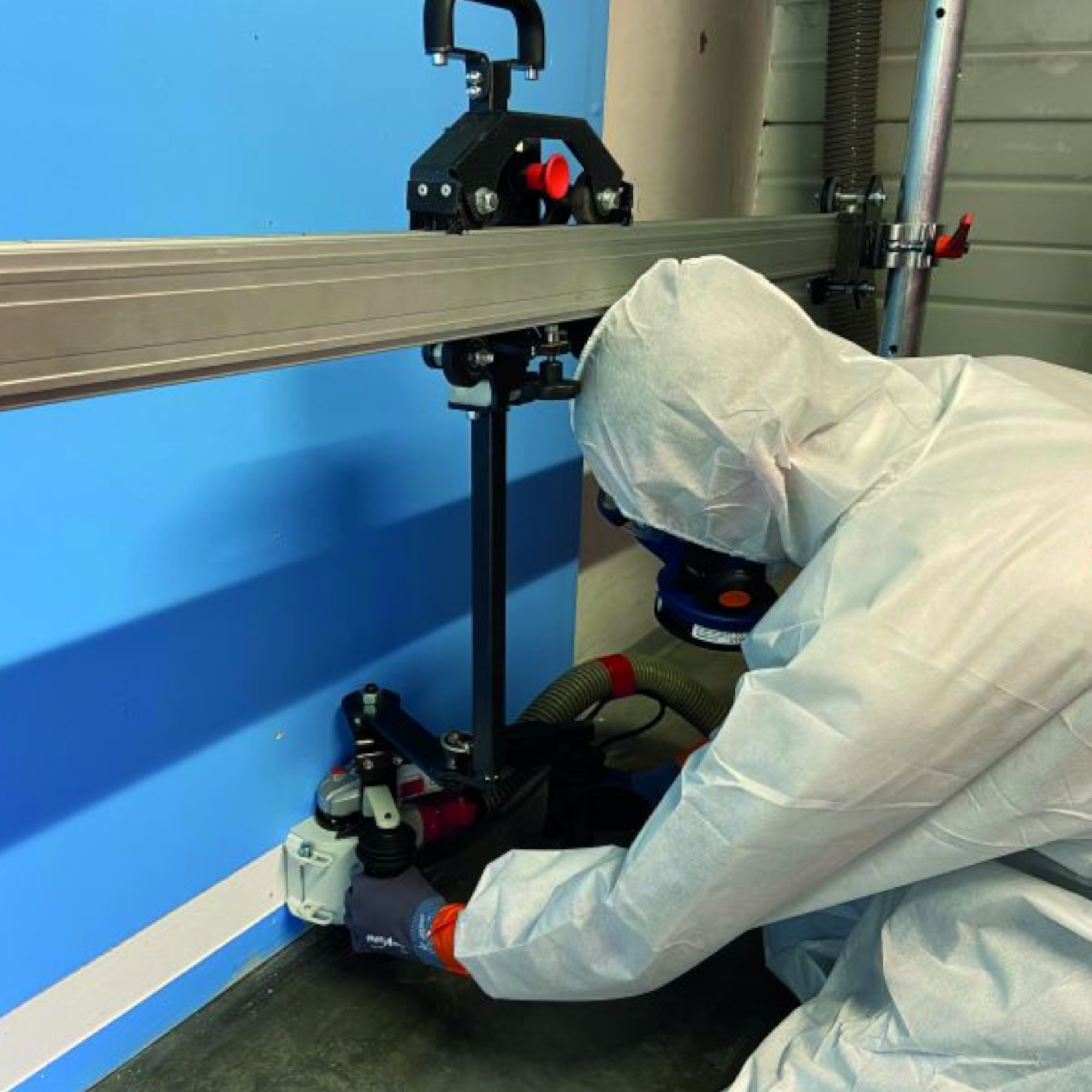 Eine Person in Schutzkleidung arbeitet an einer Maschine, die an einer Wand montiert ist. Die Maschine scheint mit einer Gleitschiene und einer Absaugvorrichtung verbunden zu sein. Der Fokus liegt auf der präzisen Handhabung des Geräts in einem industriellen Umfeld.