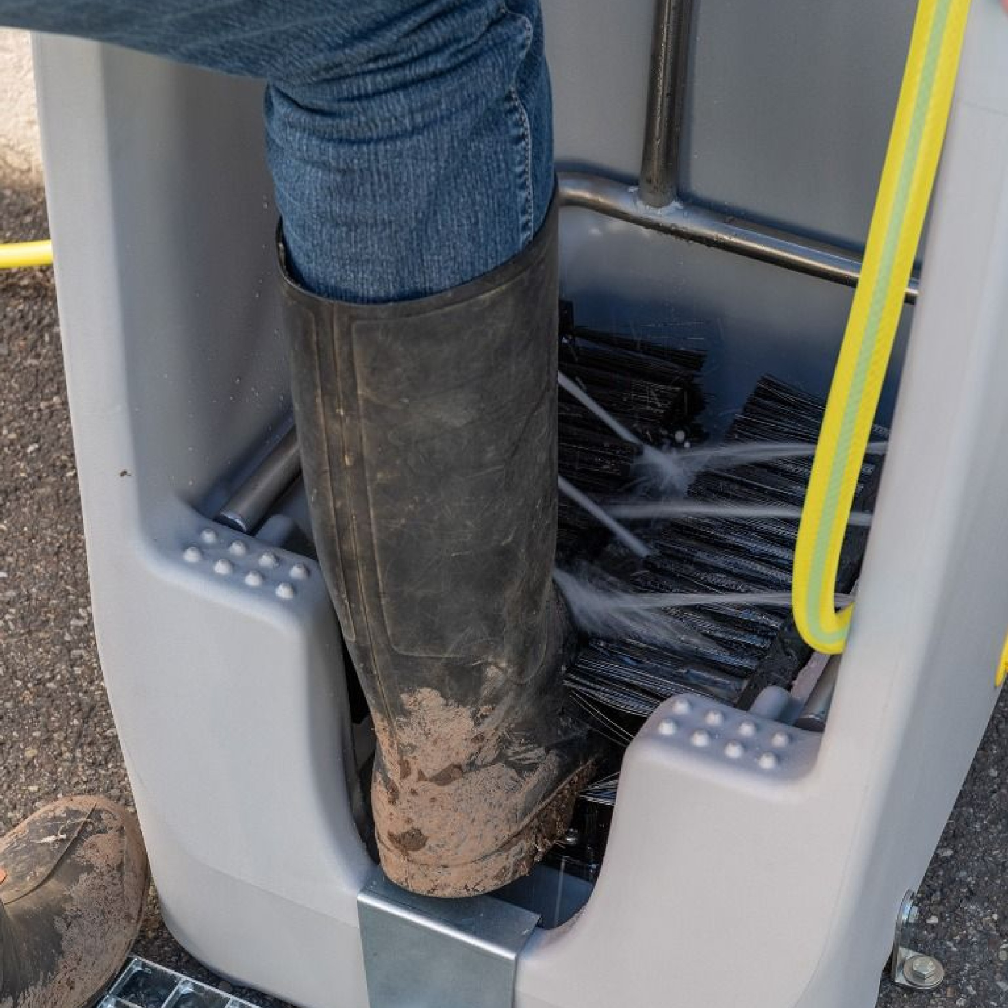 Eine Person reinigt einen schmutzigen Gummistiefel in einem speziellen Waschgerät. Das Gerät sprüht Wasser auf die Sohle, um den Schmutz zu entfernen. Die Person trägt Jeans und der Stiefel ist sichtbar verschmutzt.