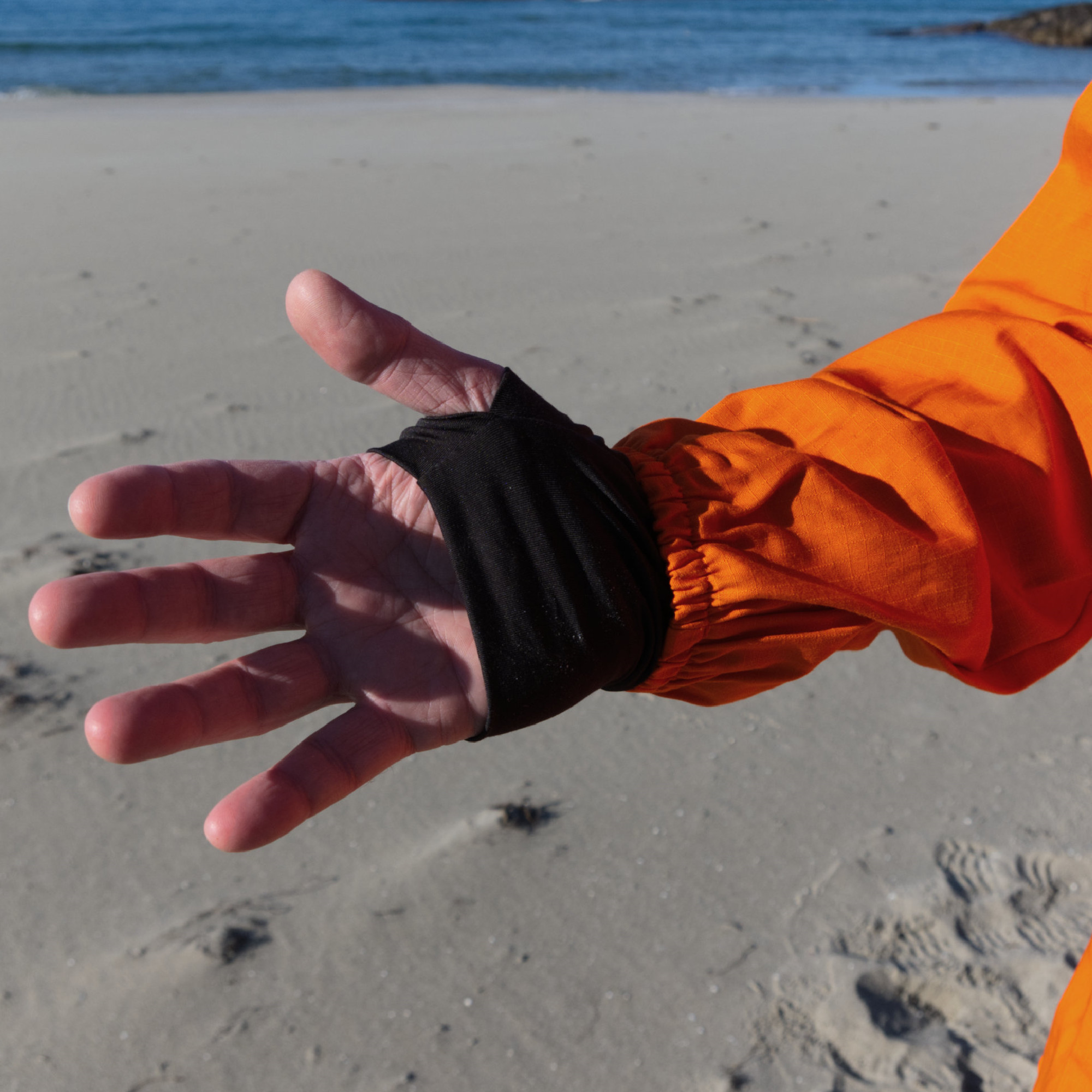 Ein ausgestreckter Hand mit einer schwarzen Manschette, die den Unterarm umschließt, vor einer sandigen Strandlandschaft. Im Hintergrund ist das Wasser sichtbar.
