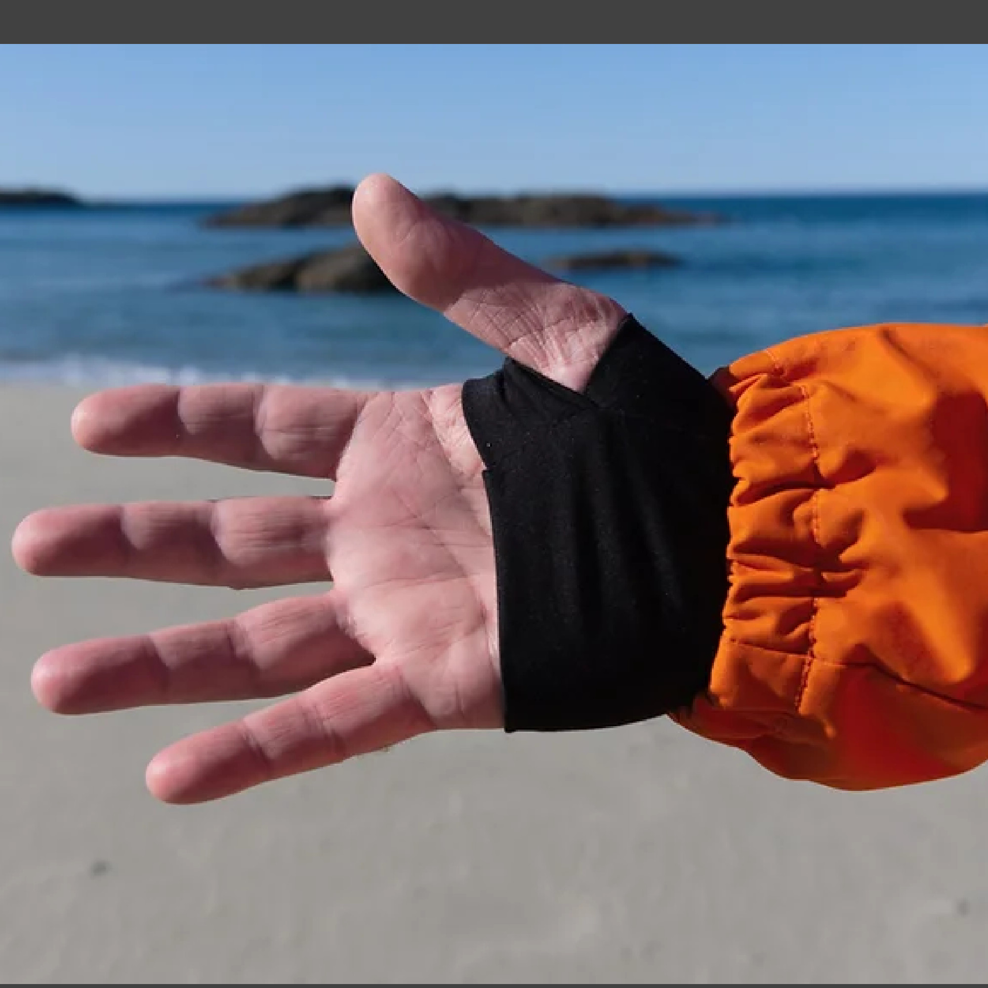 Eine offene Hand wird vor einem Strand mit Wasser im Hintergrund gezeigt. Der Handgelenkbereich ist mit einem schwarzen Band oder einer Manschette umwickelt.
