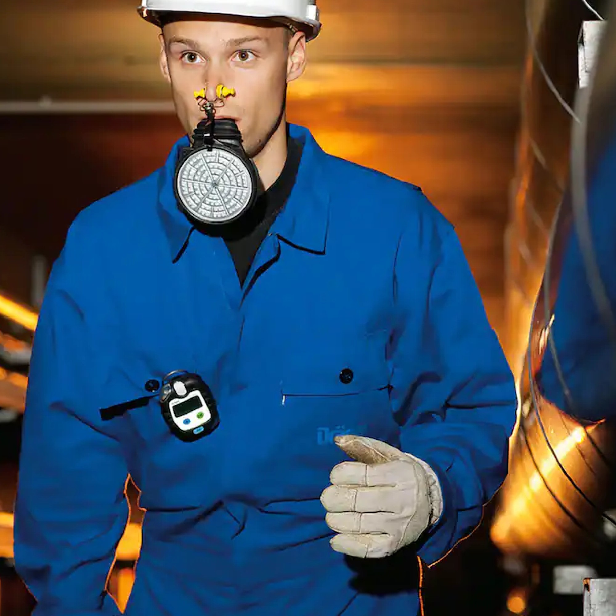 Ein junger Mann trägt einen blauen Overalls, eine Schutzmaske und einen Helm. Er hält sich an einem Objekt fest und scheint sich in einer industriellen Umgebung zu befinden. Die Beleuchtung ist gedämpft, was auf eine arbeitsintensive Situation hinweist.