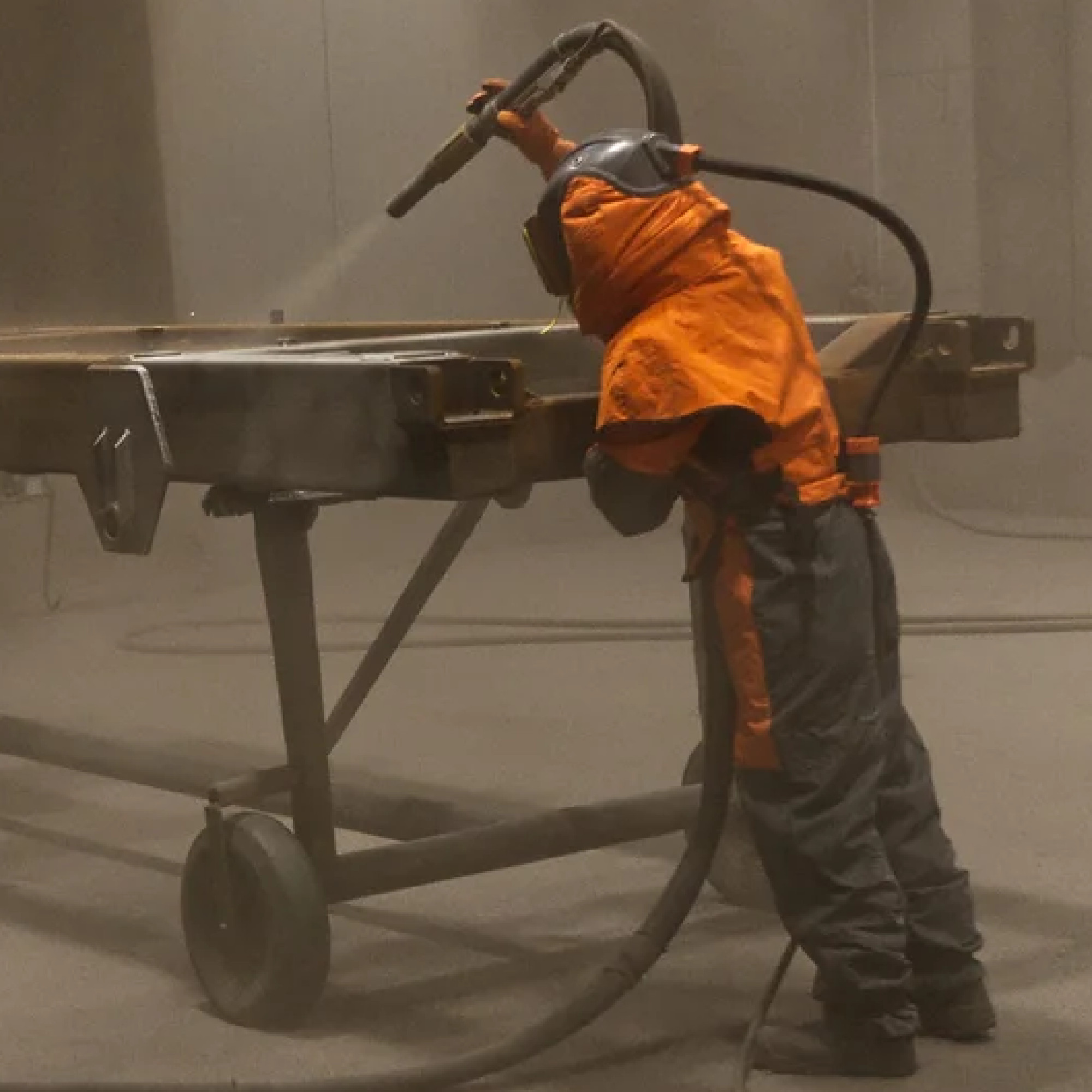 Ein Arbeiter in einem orangefarbenen Schutzanzug sprüht eine Oberfläche mit einem Schlauch in einer staubigen Umgebung. Die Szene zeigt industrielles Arbeiten, bei dem Schutzmaßnahmen getroffen werden.