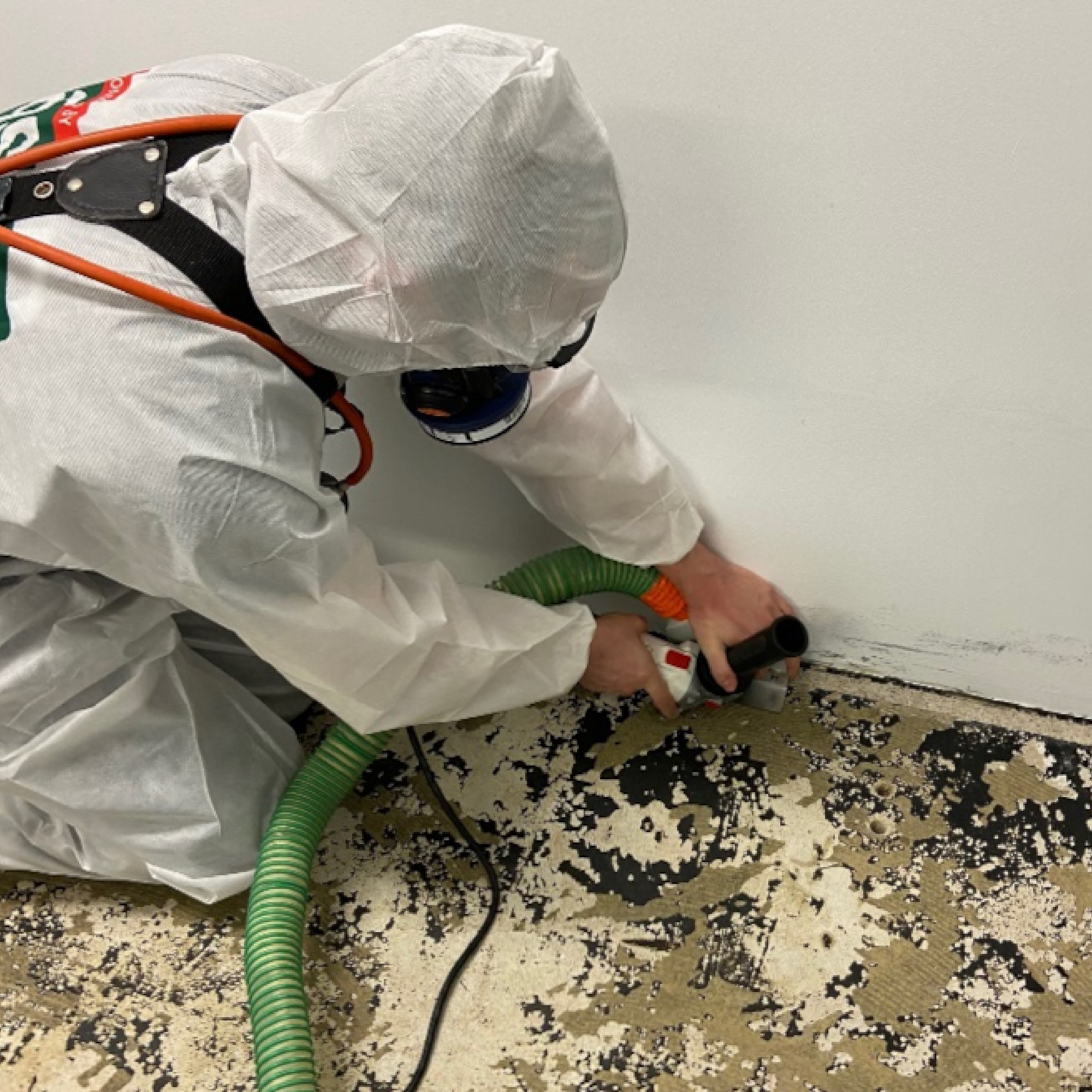 Eine Person in Schutzkleidung arbeitet am Boden. Sie hält ein Werkzeug in der Hand und benutzt einen grünen Schlauch, um den Bereich an der Wand zu reinigen oder abzudichten. Der Boden ist beschädigt und zeigt Verschmutzungen.