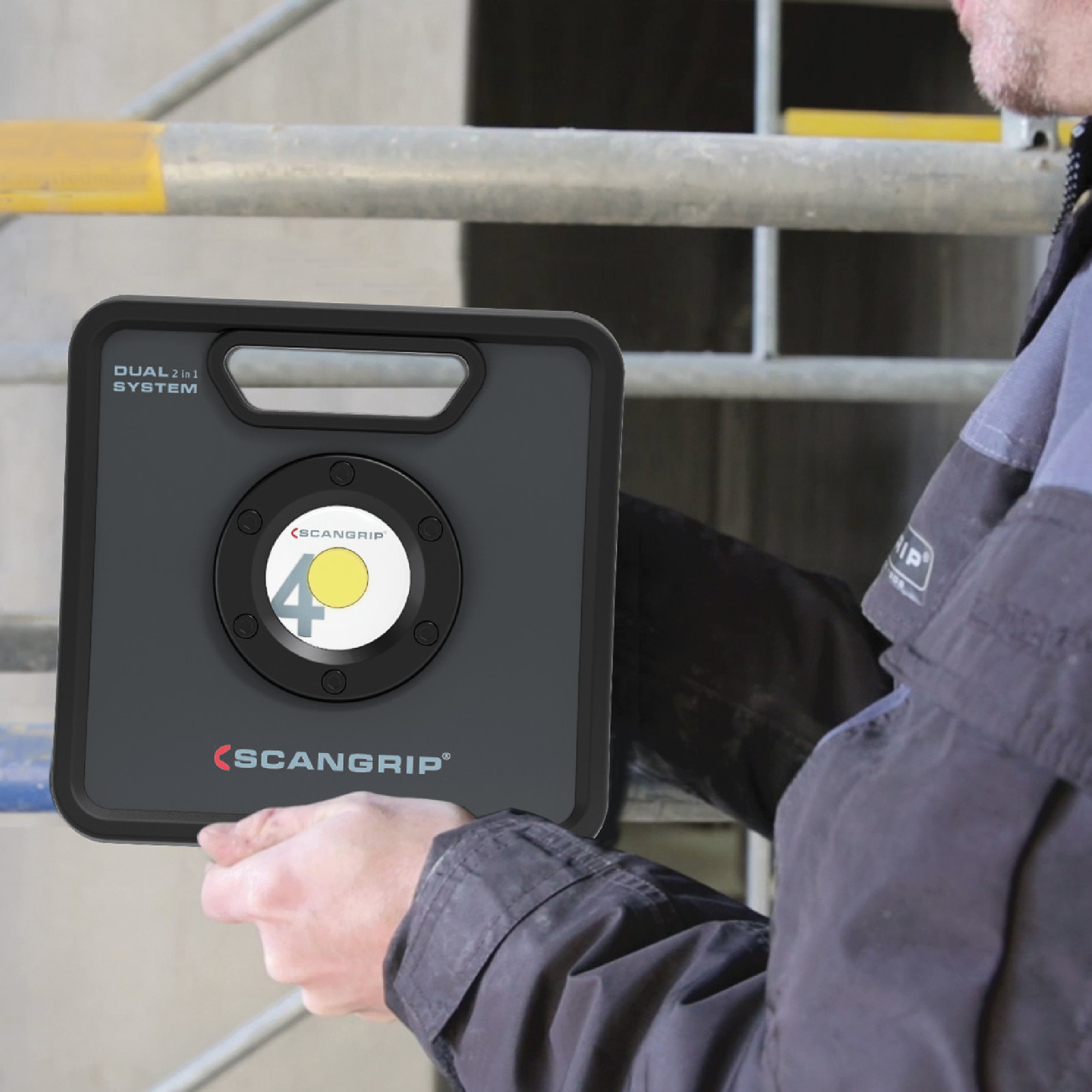Eine Person hält ein tragbares LED-Licht des Herstellers Scangrip in der Hand. Auf dem Gerät ist das Logo mit der Zahl 40 und der Aufschrift "DUAL 2 IN 1 SYSTEM" zu sehen. Der Hintergrund zeigt eine Baustelle mit Gerüsten.