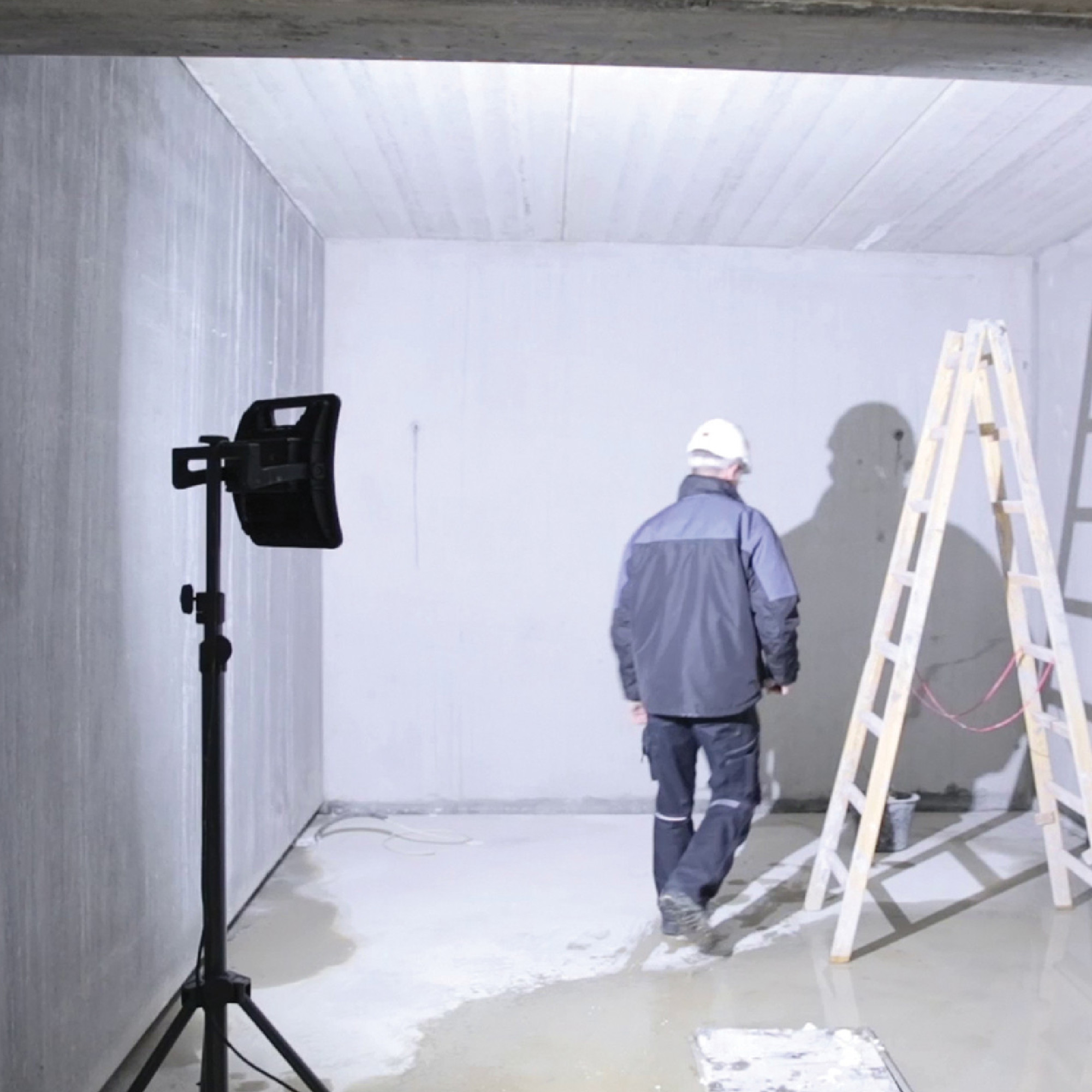 Ein Handwerker in Arbeitskleidung geht in einem leeren Raum mit grauen Wänden und frischem Estrich. Im Hintergrund steht eine Leiter und ein Baustrahler beleuchtet die Szene. Der Handwerker trägt einen weißen Helm und scheint die Umgebung zu inspizieren.