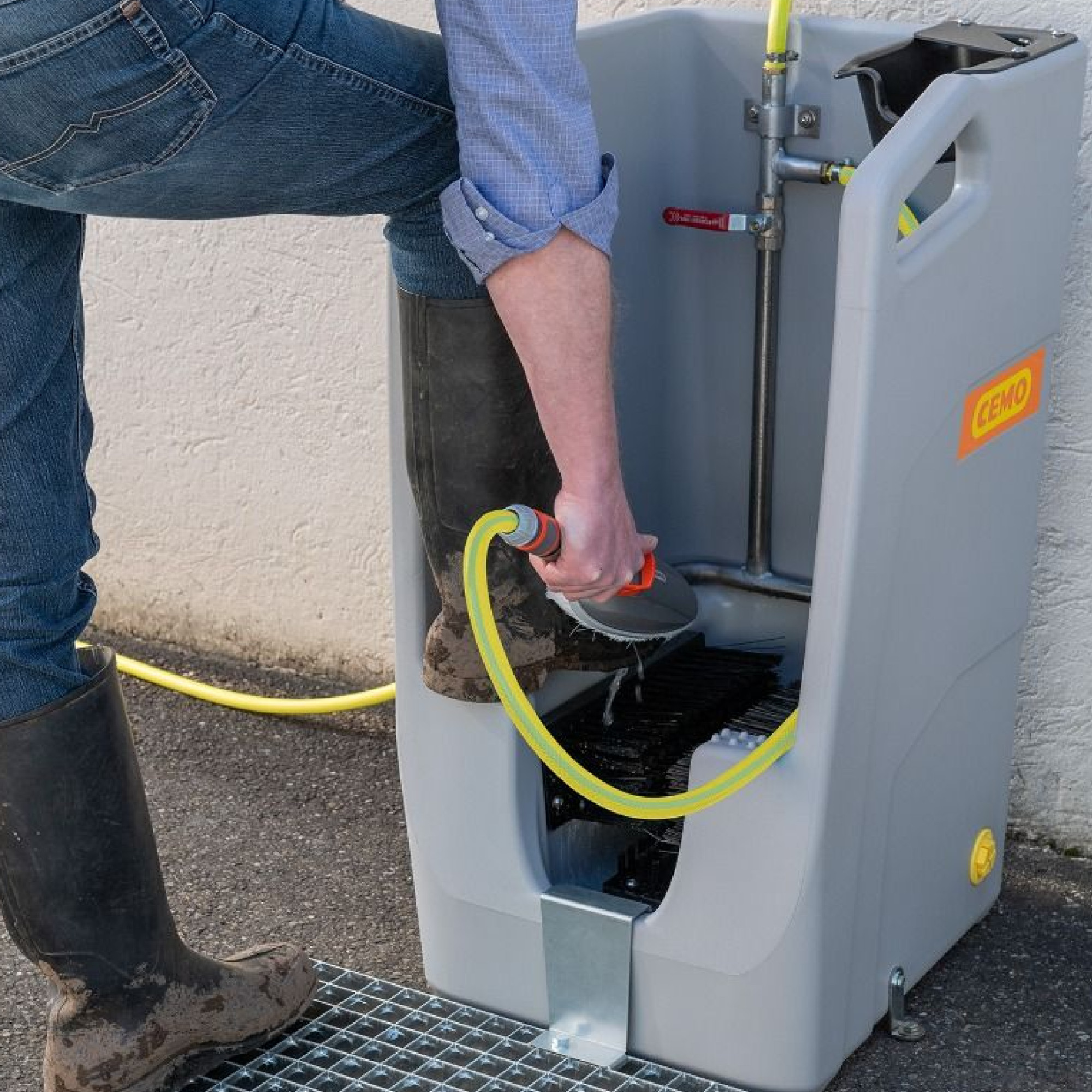 Eine Person steht mit einem Bein auf einer Reinigungsstation, die zum Reinigen von Stiefeln genutzt wird. Mit einer Hand hält sie einen Schlauch, um Schmutz von ihrem Stiefel abzuspülen. Die Station ist aus grauem Kunststoff und hat eine gelbe Schlauchverbindung.