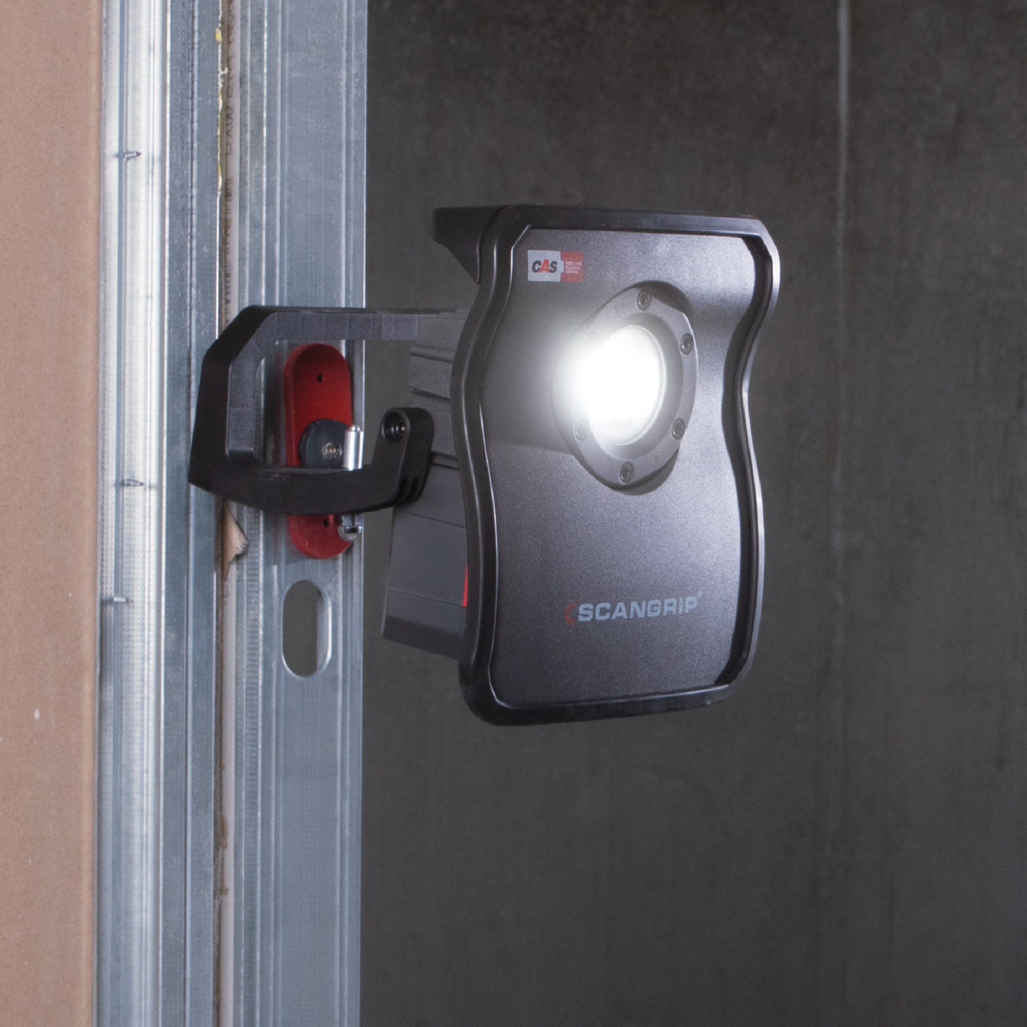 Ein schwarzer LED-Arbeitsstrahler von SCANGRIP, der an einer Wandmontage befestigt ist. Die Lampe strahlt helles Licht aus und beleuchtet den Bereich vor ihr. Das Design ist kompakt und funktional, geeignet für den Einsatz in Werkstätten oder auf Baustellen.