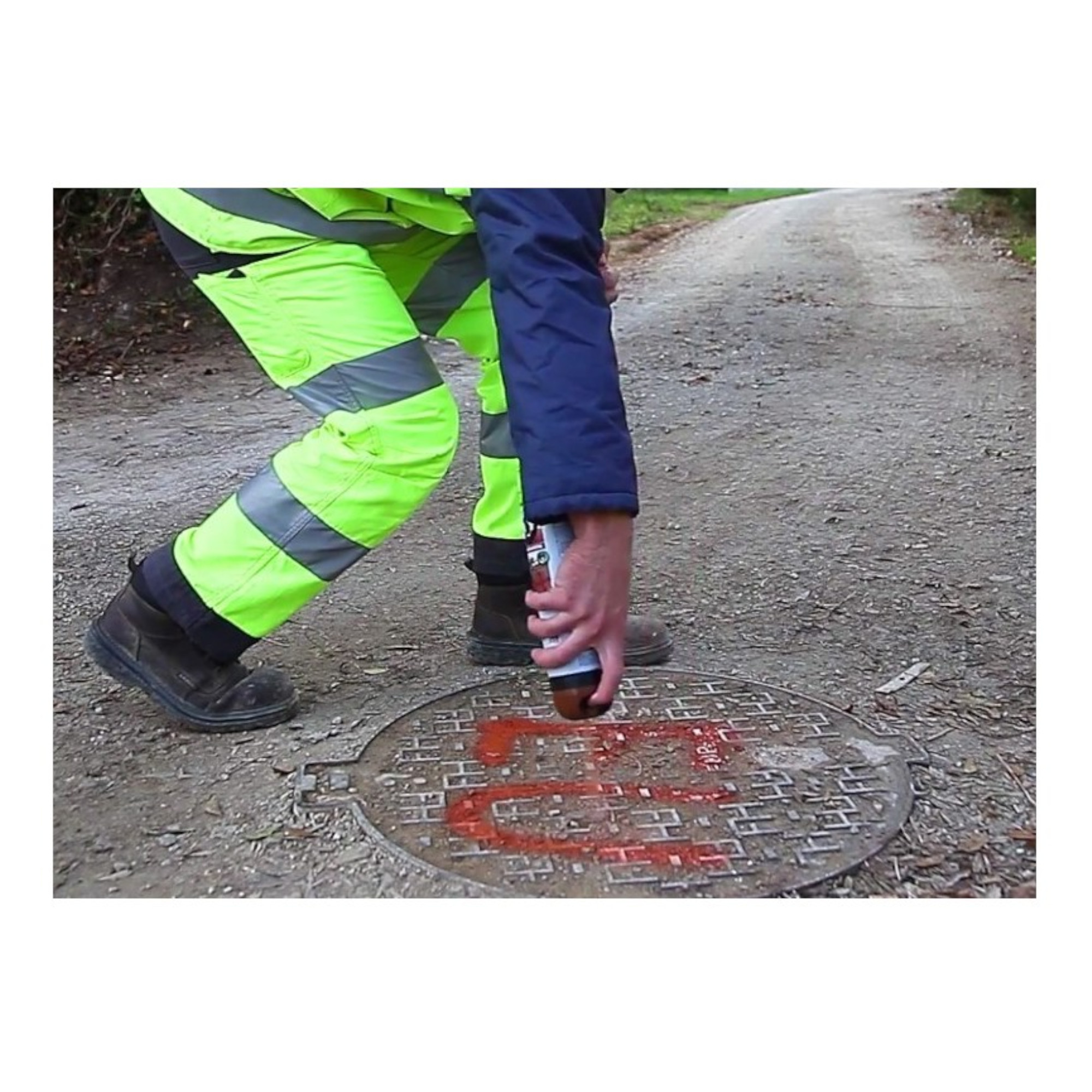 Eine Person in leuchtend gelber Arbeitskleidung sprüht mit einer Dose Farbe auf einen Gullydeckel auf einem Schotterweg. Der Gullydeckel ist rund und hat ein Muster, während der Hintergrund eine grüne, baumreiche Landschaft zeigt.