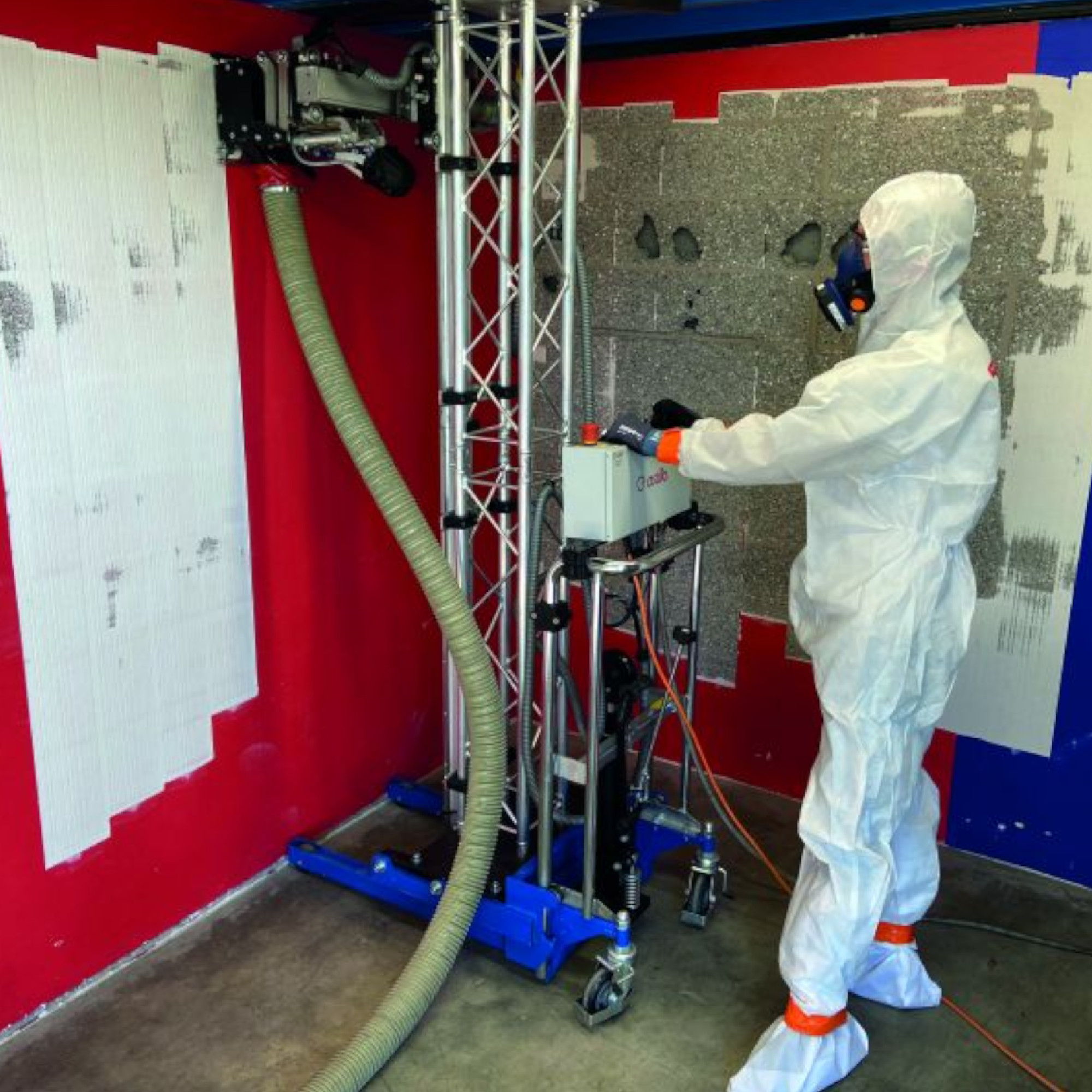 Eine Person in einem weißen Schutzanzug arbeitet mit einer Maschine in einem Raum mit roten Wänden. Die Maschine hat einen Schlauch, der zum Wandanstrich oder zur Oberflächenbearbeitung verwendet wird. частично blätternde Ecken sind sichtbar.