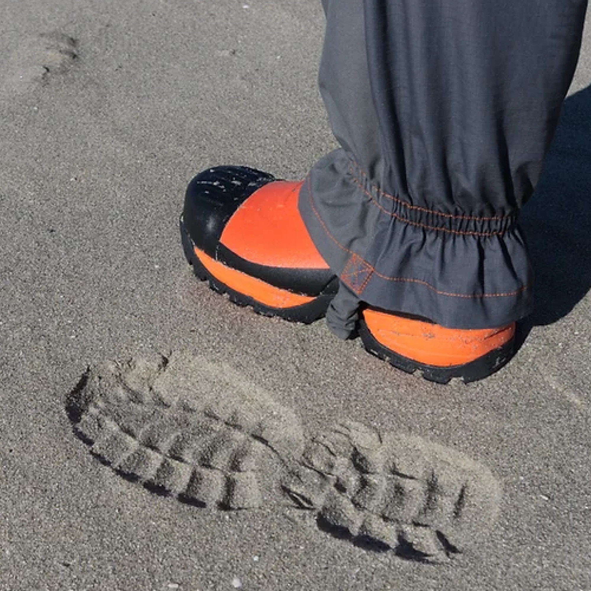 Ein Fuß mit einem orangefarbenen Schuh steht auf sandigem Boden. Neben dem Schuh sind die Fußabdrücke im Sand deutlich sichtbar. Die Hose ist am Knöchel leicht gerafft und schützt vor Schmutz.