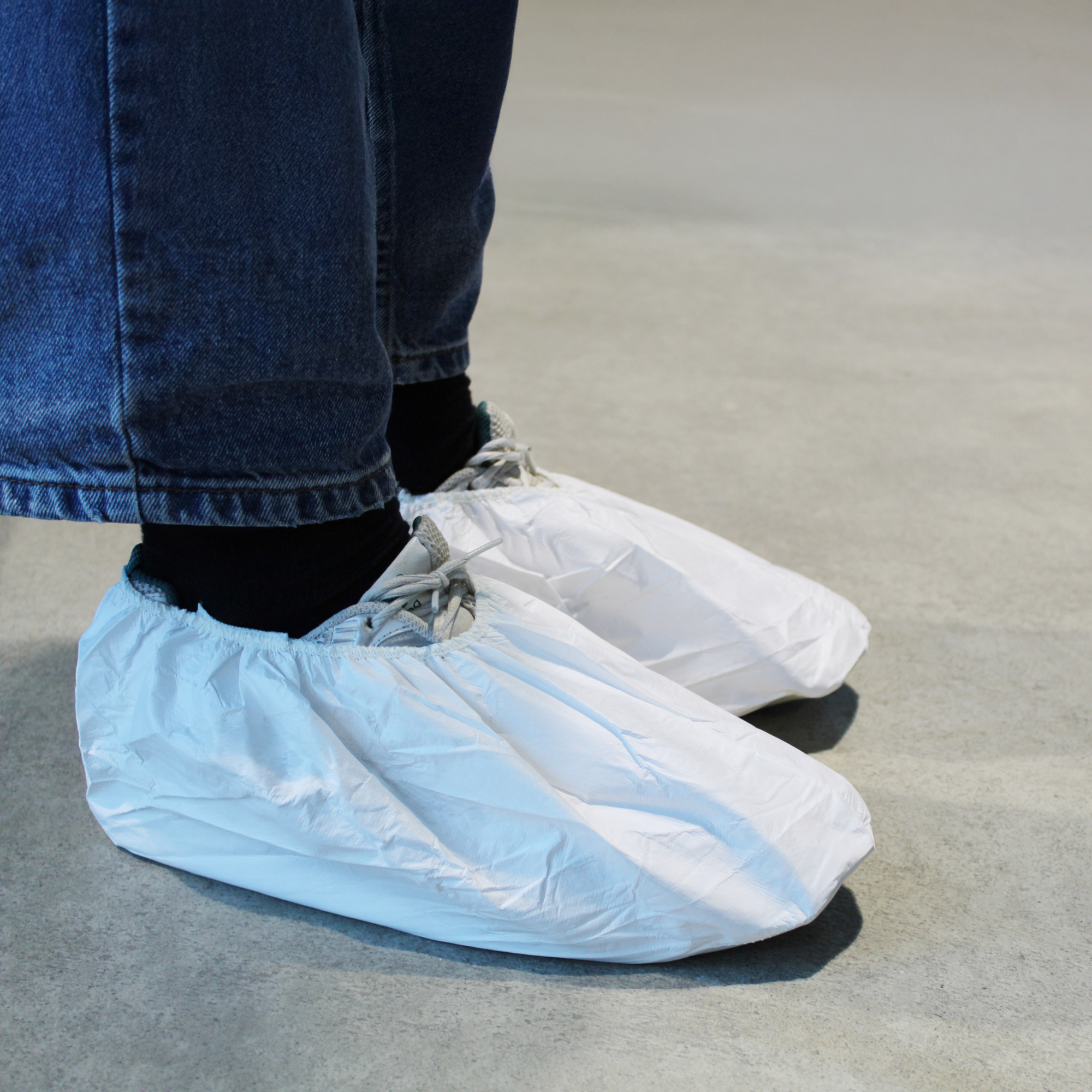 Eine Person trägt weiße Schutzüberzüge über ihren Schuhen, während sie auf einem grauen Boden steht. Die Hose ist dunkelblau, und die Unterbekleidung ist schwarz, was auf eine hygienische Umgebung hinweist.