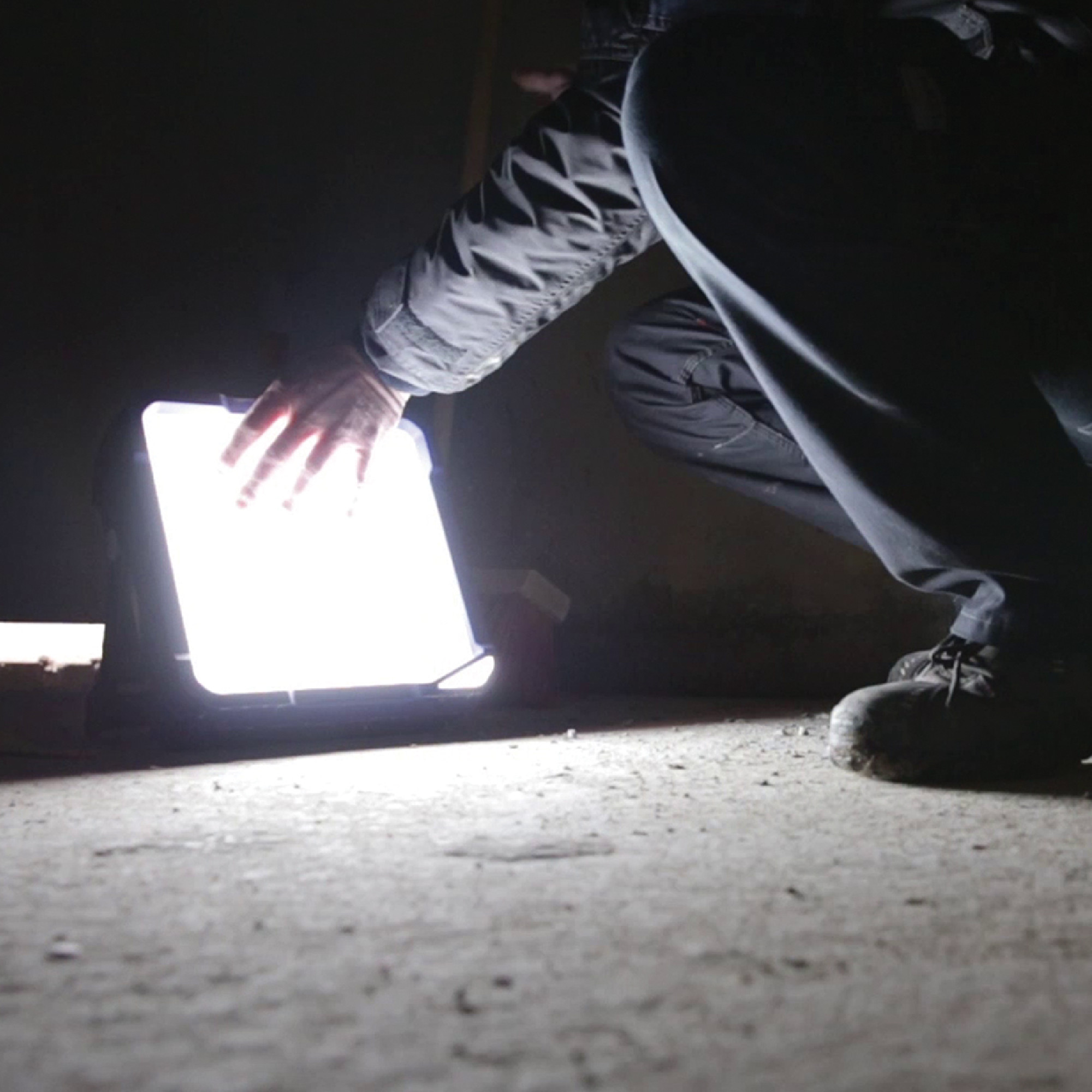 Eine Person kniet sich nieder und berührt einen mobilen LED-Scheinwerfer, der helles Licht abgibt. Die Umgebung ist dunkel und der Scheinwerfer beleuchtet den Boden in der Nähe.