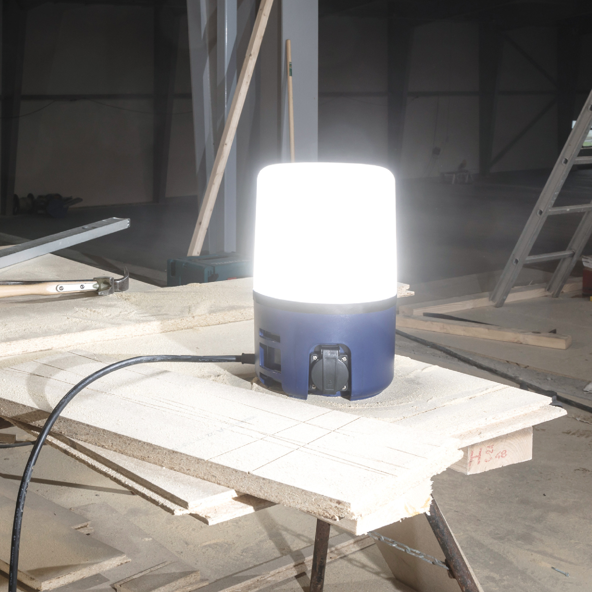 Eine helle, runde Baustellenlampe steht auf einer Tischplatte aus Holz. Die Lampe strahlt starkes Licht aus und befindet sich in einem Baustellenumfeld mit Holzplatten und Werkzeugen im Hintergrund.