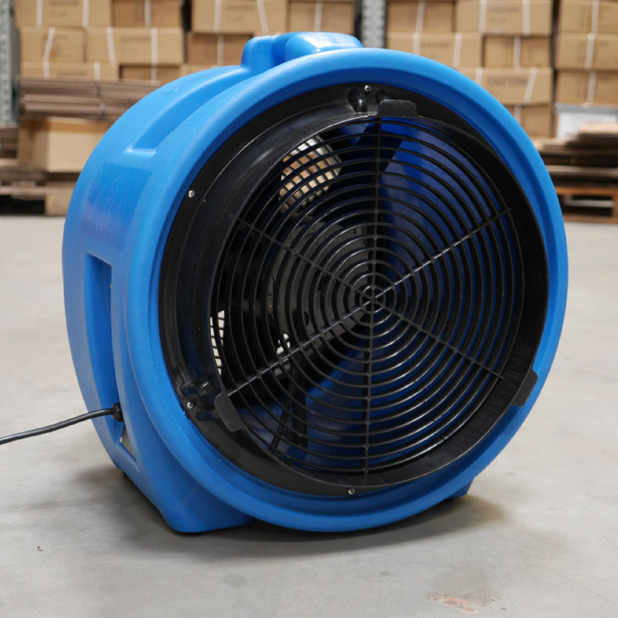 Ein blauer Ventilator steht auf einem Betonboden. Er hat ein rundes Gehäuse und ein schwarzes Gitter vor dem Rotor. Der Ventilator ist darauf ausgelegt, Luft zu bewegen und könnte in industriellen oder gewerblichen Umgebungen verwendet werden.