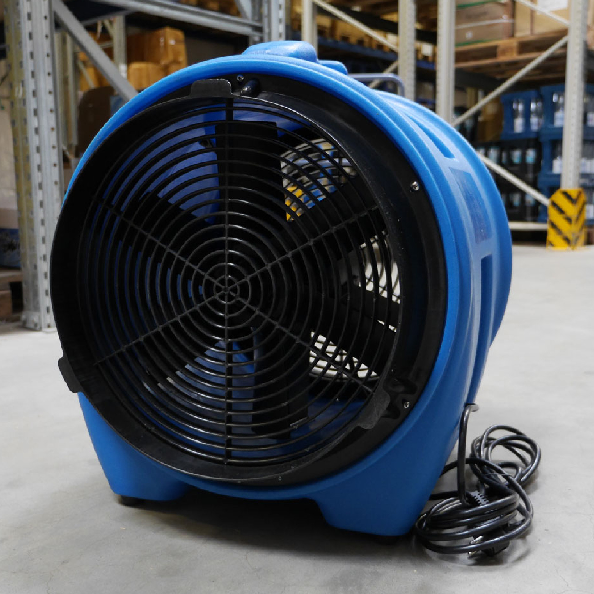 Ein blauer Ventilator steht auf einem Betonboden. Die Vorderseite zeigt das runde Gitter der Lüfterblätter. Im Hintergrund sind Regale mit Kisten und Verpackungen zu sehen, die auf eine Lagereinrichtung hinweisen.