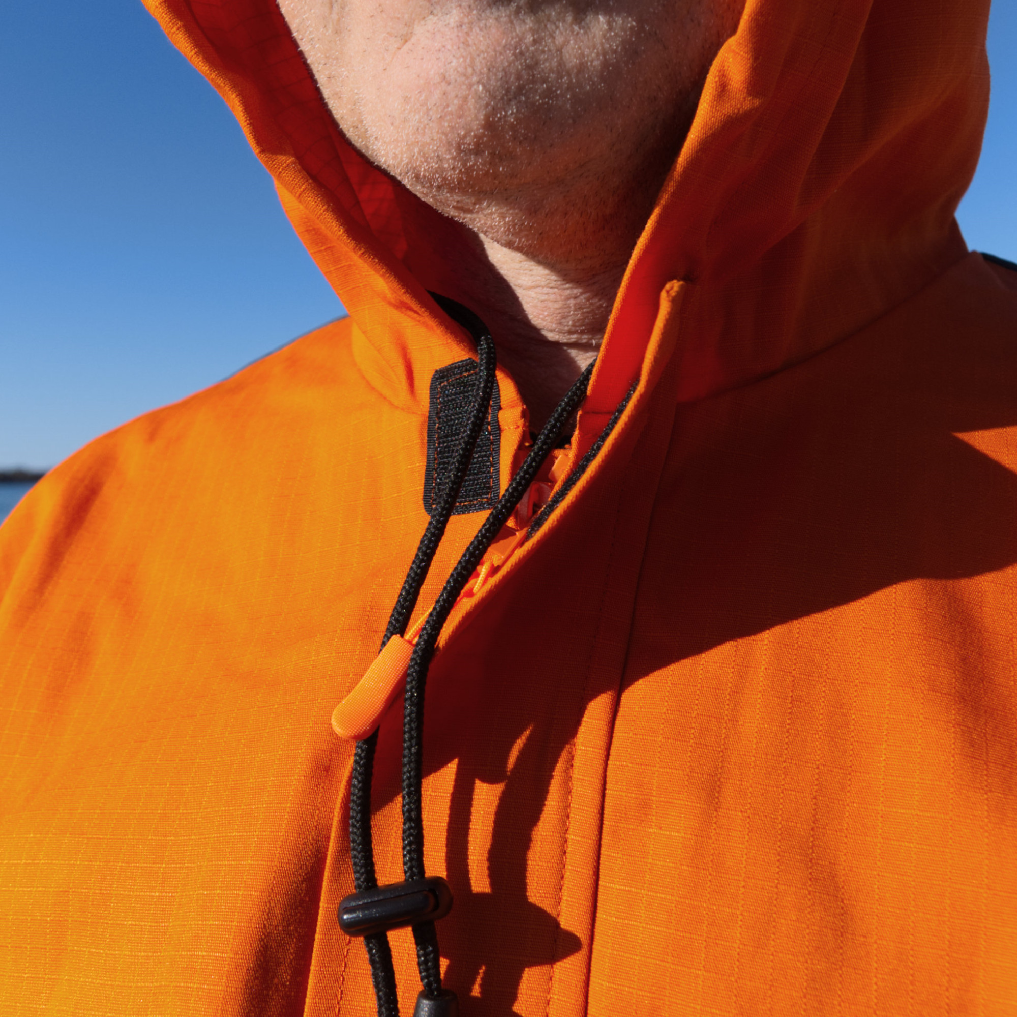 Nahaufnahme einer Person in orangefarbener Jacke mit Kapuze. Die Jacke hat schwarze Kordeln am Hals. Der Hintergrund zeigt einen klaren blauen Himmel und einen unscharfen Wasserbereich. Das Bild vermittelt eine Outdoor-Atmosphäre.